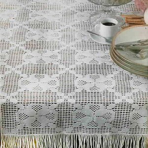 May include: A white, crocheted tablecloth with a geometric pattern and fringe. A glass bowl filled with fruit sits on the table, along with a cup of coffee and a stack of plates. The tablecloth is likely 60 inches wide.