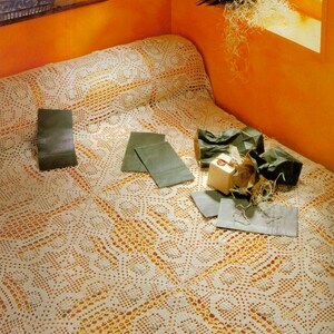 May include: A cream-colored, crocheted bedspread with a detailed, openwork pattern. Several small, gray paper bags and a small gift box are arranged on the bedspread. The room has orange walls and a window.