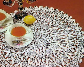 Può includere: Una tovaglietta rotonda bianca all'uncinetto con un motivo floreale dettagliato. Due tazze da tè con disegni floreali sono sulla tovaglietta, insieme a un limone e un candelabro d'argento con candele rosse. Lo sfondo è marrone pieno.