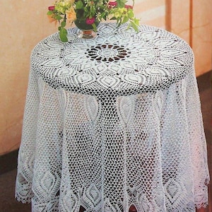 May include: A white, round, crocheted tablecloth covers a small table. The intricate lace design features a central floral pattern and a scalloped edge. A vase of colorful flowers sits on top of the table, adding a touch of color.
