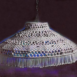 Puede incluir: Una lámpara colgante de macramé color crema. La pantalla tiene una forma cónica ancha con un diseño de tejido abierto y un ribete de flecos. La luz cuelga de una cadena.