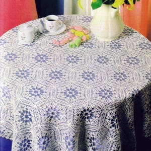May include: A round, white crocheted tablecloth with a floral pattern covers a table. A white sugar bowl, a cup and saucer with a rose design, and a white vase with yellow flowers are on the table. A few candies are also present.