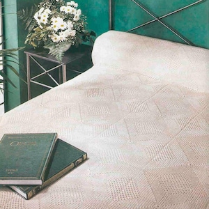 May include: A cream-colored bedspread featuring a repeating diamond pattern with intricate knit textures. The textile covers a bed surface, and two dark green hardcover books with gold lettering are placed on the lower left corner of the fabric.