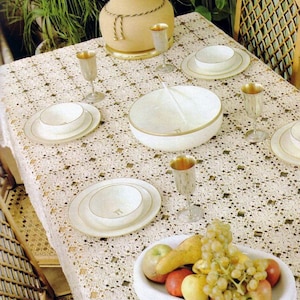 Könnte beinhalten: Ein Esstisch mit einer cremefarbenen gehäkelten Tischdecke, weißen Keramiktellern und Schalen mit Goldrand, silberfarbenen Kelchen und einer weißen ovalen Servierschale, die mit verschiedenen frischen Früchten wie grünen Trauben und Äpfeln gefüllt ist.