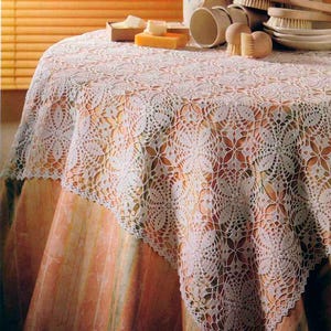 May include: A round table covered with a white, intricately crocheted lace tablecloth. The table is set with various bath accessories, including soap, brushes, and containers. The background features a window with orange blinds.