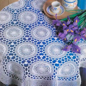 May include: A white round crochet tablecloth featuring a repeating circular motif pattern. The tablecloth is draped over a surface, with a wooden tray holding a ceramic cup, a small lidded sugar bowl, and a bouquet of purple iris flowers.