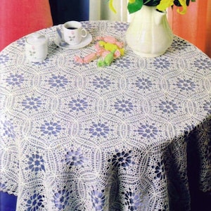 May include: A round white crochet tablecloth featuring a repeating floral and geometric pattern. On the table sits a white ceramic pitcher with yellow lilies, a white ceramic sugar bowl, a white teacup with a rose motif, and colourful sweets.