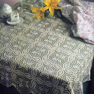 May include: A crocheted tablecloth with a delicate floral pattern in a cream colour. The tablecloth is laid on a table with three glasses of water and yellow flowers.