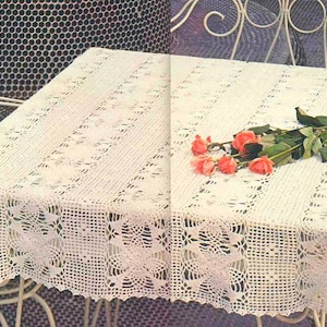May include: A rectangular off-white crochet tablecloth featuring an intricate openwork pattern with repeating geometric and floral motifs. A small bouquet of five salmon-colored roses with green stems and leaves rests on the fabric surface.