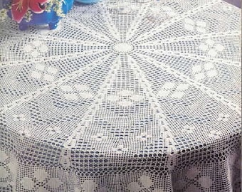 May include: White crocheted tablecloth with a delicate lace pattern. The tablecloth is round and has a center design with a circular motif. The tablecloth is laid on a table with a dark surface.