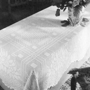 May include: A white, intricately crocheted tablecloth with a geometric pattern covers a table. The tablecloth has a scalloped edge and is partially covering the table. A vase with flowers sits on the table.