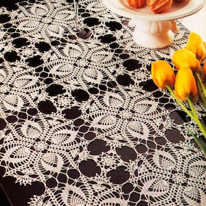 May include: White crocheted table runner with a delicate floral pattern. The runner is laid on a black surface with a glass of wine and a plate of fruit on a white pedestal.