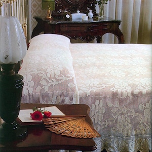 May include: A white crocheted bedspread with a floral pattern. The bedspread is on a bed with a white pillow and a wooden headboard. A wooden table with a lamp and a fan is in the foreground.