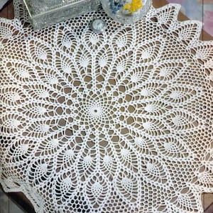 May include: A white, intricately crocheted doily with a circular design. The doily is placed on a wooden surface, with a silver box, a candle, and a small clock visible in the background. The doily's pattern is symmetrical and detailed.