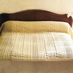 May include: A cream-colored knitted bedspread featuring a complex pattern of vertical cable stitches, braided textures, and zigzag motifs. The textile covers the surface of a bed, displaying a uniform off-white hue and a dense, tactile knit construction.