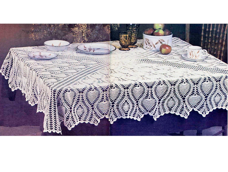 May include: White crocheted tablecloth with a delicate lace pattern and a scalloped edge. The tablecloth is laid on a table with a purple surface and features a central design of repeating hearts.