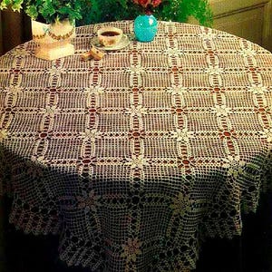 May include: A round, cream-colored crocheted tablecloth with a detailed openwork pattern. The tablecloth is draped over a dark surface, with a cup of coffee, a small plate, and a vase of flowers visible on the table.