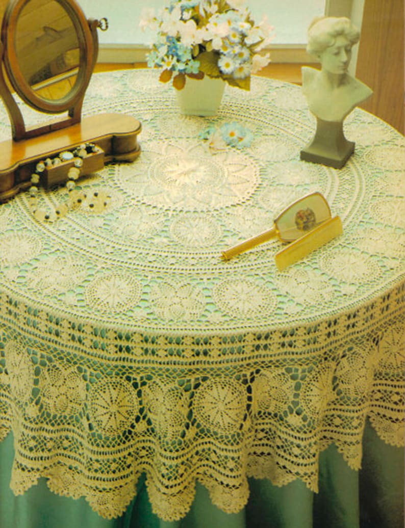 May include: A crocheted tablecloth with a delicate lace pattern in a cream color. The tablecloth is laid on a green table and features a round design with a scalloped edge.