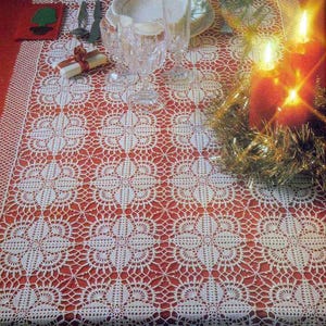 Puede incluir: Una mesa festiva con un mantel rojo con un patrón floral blanco tejido a ganchillo. La mesa está puesta con copas de cristal, un plato, cubiertos y velas encendidas, creando un ambiente cálido y acogedor para una comida festiva.