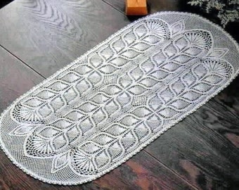 Può includere: Un runner da tavolo bianco all'uncinetto con un delicato e intricato motivo di foglie e volute. Il runner è di forma ovale e presenta un design traforato e in pizzo.