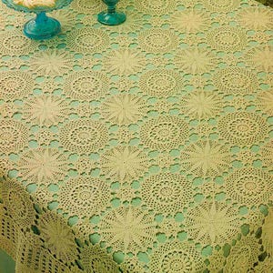 May include: A cream-colored, crocheted tablecloth with a repeating floral pattern. The tablecloth is draped over a table, with a turquoise glass dish and a turquoise glass goblet visible on the table. The tablecloth's edges have a decorative border.