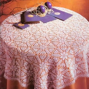 May include: A round white crochet tablecloth with an intricate floral pattern draped over a table. On top of the tablecloth are two dark purple spherical candles, several purple rectangular cards with circular gold accents, and straw.