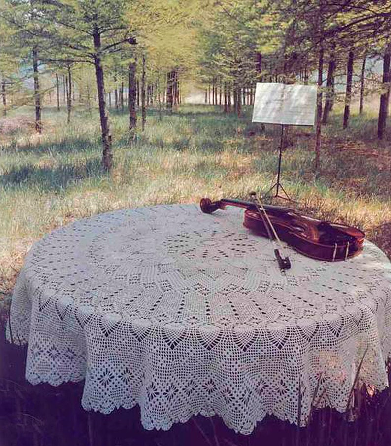 May include: A white crocheted tablecloth with a delicate floral pattern covers a round table. A violin and bow are placed on the tablecloth. The table is set in a forest clearing with a music stand in the background.