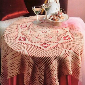 以下が含まれることがあります： クリーム色の繊細な花柄のクロシェのテーブルクロス。テーブルクロスは赤いテーブルに置かれ、ワイングラスが2つとチョコレートの入った皿がスタンドの上に置かれています。