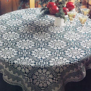 May include: A round white crochet tablecloth featuring an intricate floral and geometric pattern with a scalloped edge, draped over a circular table. The lace fabric displays a detailed openwork design with repeating motifs across the entire surface.