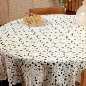 以下が含まれることがあります： 繊細な花柄の白いクロシェのテーブルクロス。テーブルクロスは、中央にぶどうの入ったボウルのある木製のテーブルの上に置かれています。