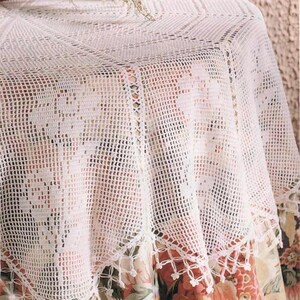 May include: White crocheted table cloth with a delicate floral pattern and a fringe border. The tablecloth is laid on a table with a floral print tablecloth.
