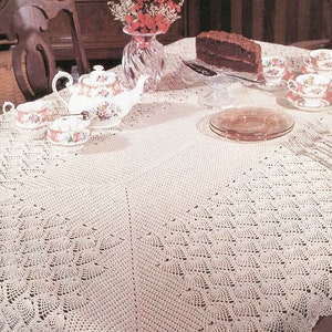 Könnte beinhalten: Eine weiße Häkeltischdecke mit einem zarten Spitzenmuster, gedeckt für eine Teestunde mit Teekanne, Tassen und einem Teller Kuchen.