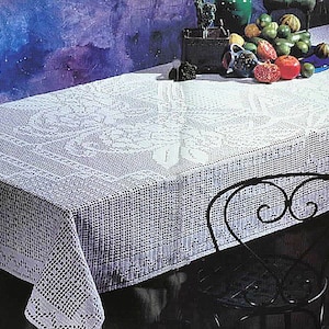 Puede incluir: Un mantel de crochet blanco con un delicado patrón floral, drapeado sobre una mesa con una silla de hierro forjado negro. El mantel tiene un borde festoneado y está hecho con un hilo fino.