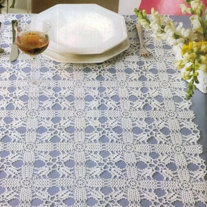 May include: A white, crocheted table runner with a delicate, openwork pattern. The runner is placed on a light blue surface and is decorated with a scalloped edge. A wine glass and plates are set on the table.