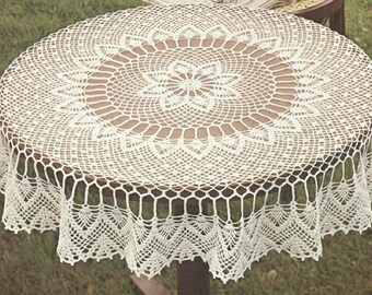 Puede incluir: Un mantel de mesa de crochet blanco con un delicado diseño floral. El mantel tiene un borde festoneado y está colocado sobre una mesa de madera.