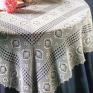 May include: Cream-colored, crocheted tablecloth with a detailed floral pattern. The round tablecloth is draped over a table, with a dark blue background. Pink flowers and a book are visible on the table.