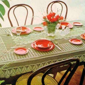 May include: A rectangular dining table covered with a light green lace tablecloth featuring a leaf-patterned border. The table is set with bright orange plates and bowls, clear glass tumblers, silver cutlery, and a glass vase filled with red flowers.