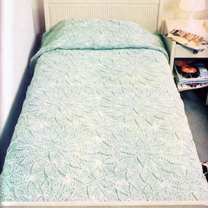 May include: A light mint green crochet bedspread with a repeating floral pattern covering a single bed. The bed features a white slatted headboard and footboard, positioned next to a white nightstand with a lamp and magazines.