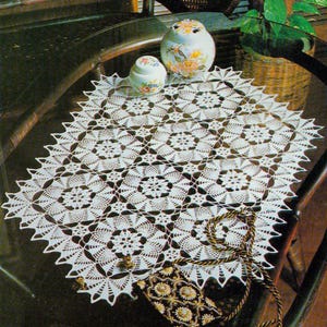 Op de afbeelding: Een witte, gehaakte doily met een bloemenpatroon, geplaatst op een glazen tafel. De doily heeft een geschulpte rand en is omgeven door decoratieve items, waaronder een kleine tas met een gouden band en twee keramische potten met bloemmotieven.