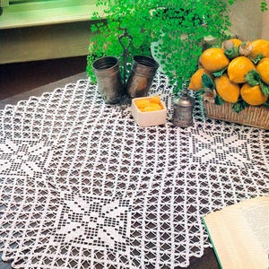 May include: A white crocheted tablecloth featuring a repeating diamond lattice pattern with square floral motifs at the corners. The textile is crafted with fine thread and displays a delicate openwork design that covers the surface of the table.
