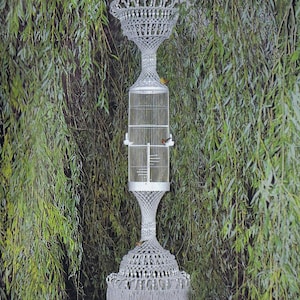 May include: A tall, white macramé birdcage stand featuring intricate knotted patterns, a central cylindrical wire cage, and a wide, fringed base. The text Macramé Enchantment appears at the top, and the word Paradise is printed at the bottom.