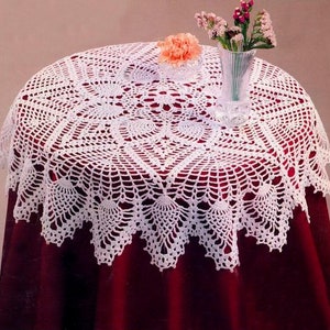 May include: White crocheted table runner with a delicate floral pattern. The runner is laid on a red tablecloth and features a scalloped edge.