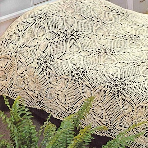 May include: A cream-colored, crocheted bedspread with an intricate, openwork design. The pattern features repeating floral and geometric shapes, creating a delicate and textured appearance. The bedspread is draped over a bed, with a glimpse of a dark surface underneath.