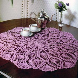 May include: A lavender crochet doily with an oval shape and intricate openwork pattern. Two teacups and saucers are placed on the doily, with a silver teapot and a vase of purple flowers. The doily's design includes a scalloped edge.