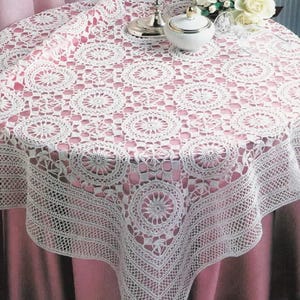 May include: A white, crocheted tablecloth with a circular pattern covers a round table. The intricate lace design features repeating floral motifs and geometric shapes. The tablecloth is draped over a pink surface.
