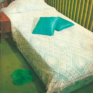 May include: A single bed dressed in a white, intricate, diamond-patterned crochet bedspread with a scalloped fringe edge. Two solid teal square throw pillows rest on the bed, and a pair of green slippers are placed on the floor beside the bed.