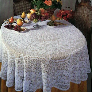 Puede incluir: Un mantel de crochet blanco con un delicado patrón floral cubre una mesa redonda. La mesa está puesta con fruta y flores, creando un centro de mesa encantador y elegante.