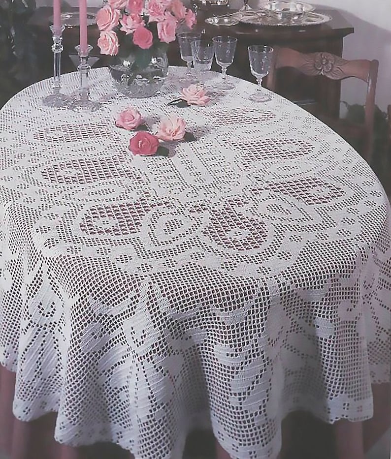 May include: White crocheted oval tablecloth with a delicate floral pattern. The tablecloth is laid on a table with a pink tablecloth underneath. There are pink roses in a vase on the table.