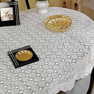 May include: White crocheted tablecloth with a delicate pattern, featuring a gold bowl and a black tray with a gold plate on a round table.