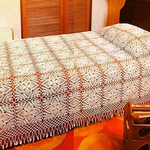 Op de afbeelding: Een crèmekleurig, gehaakt sprei met een geometrisch patroon bedekt een bed. Het sprei heeft een gefranjerde rand en een bijpassende kussensloop. Het bed staat tegen een houten hoofdbord en een houten kast.